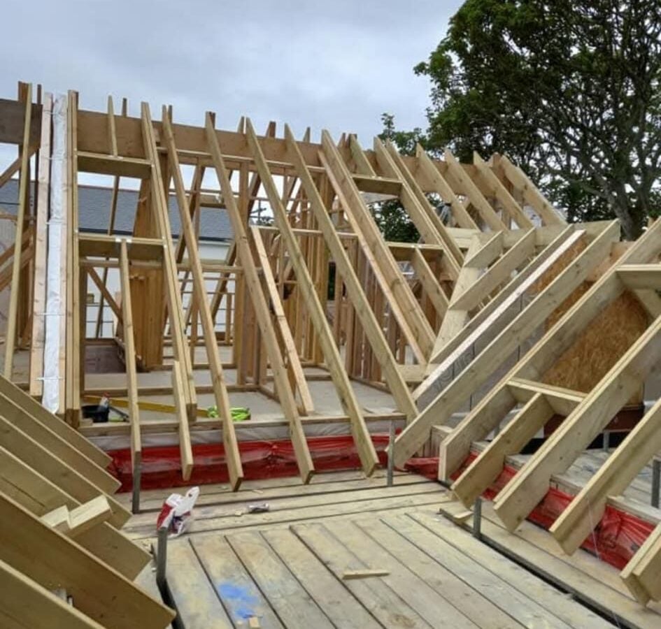 New timber roof structure under construction on a residential property, showing exposed rafters and trusses before covering materials are applied, with scaffolding and safety barriers around the site